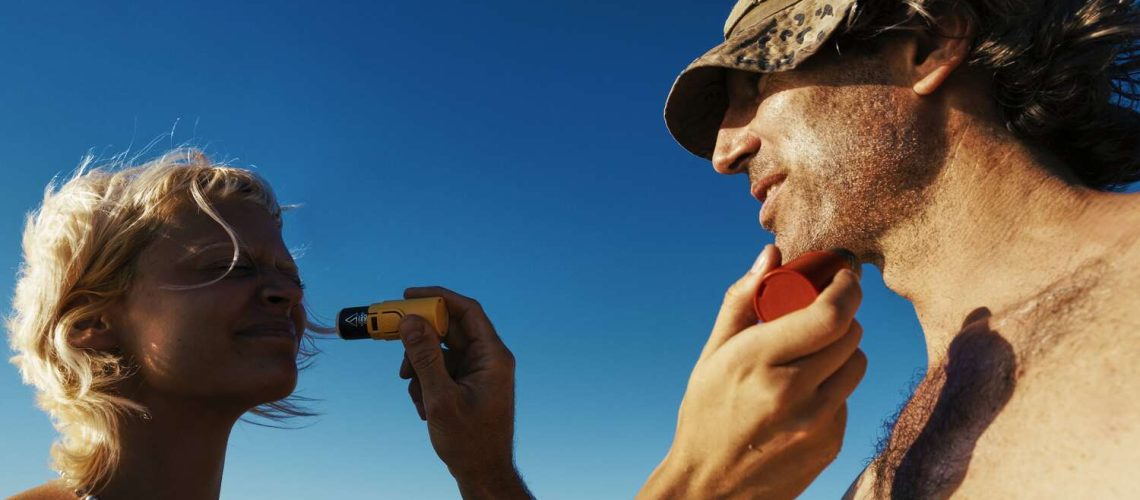 mejor-proteccion-solar-facial Una persona aplica crema solar resistente al agua en el rostro de otra persona mientras está de pie al aire libre bajo un cielo azul claro.